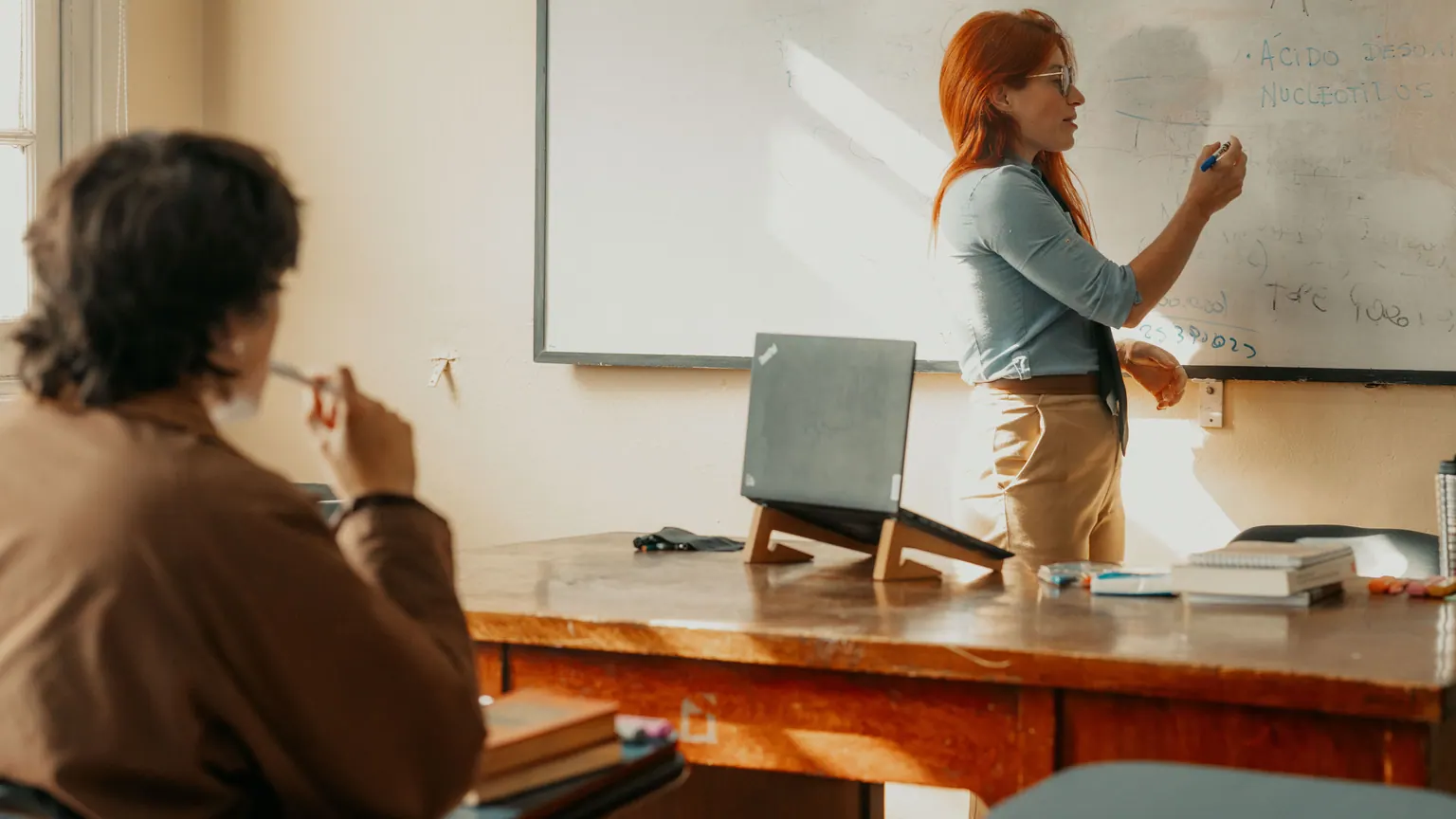 Professora escreve no quadro enquanto estudantes acompanham a atividade