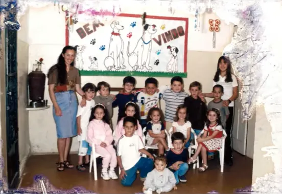 Fotografia antiga da turma da escolinha, com a professora e as crianças em frente ao mural de boas-vindas.